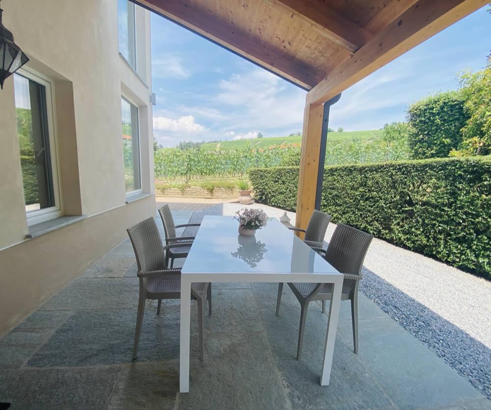 Terrazza coperta con tavolo bianco, sedie e vista su giardino verde.