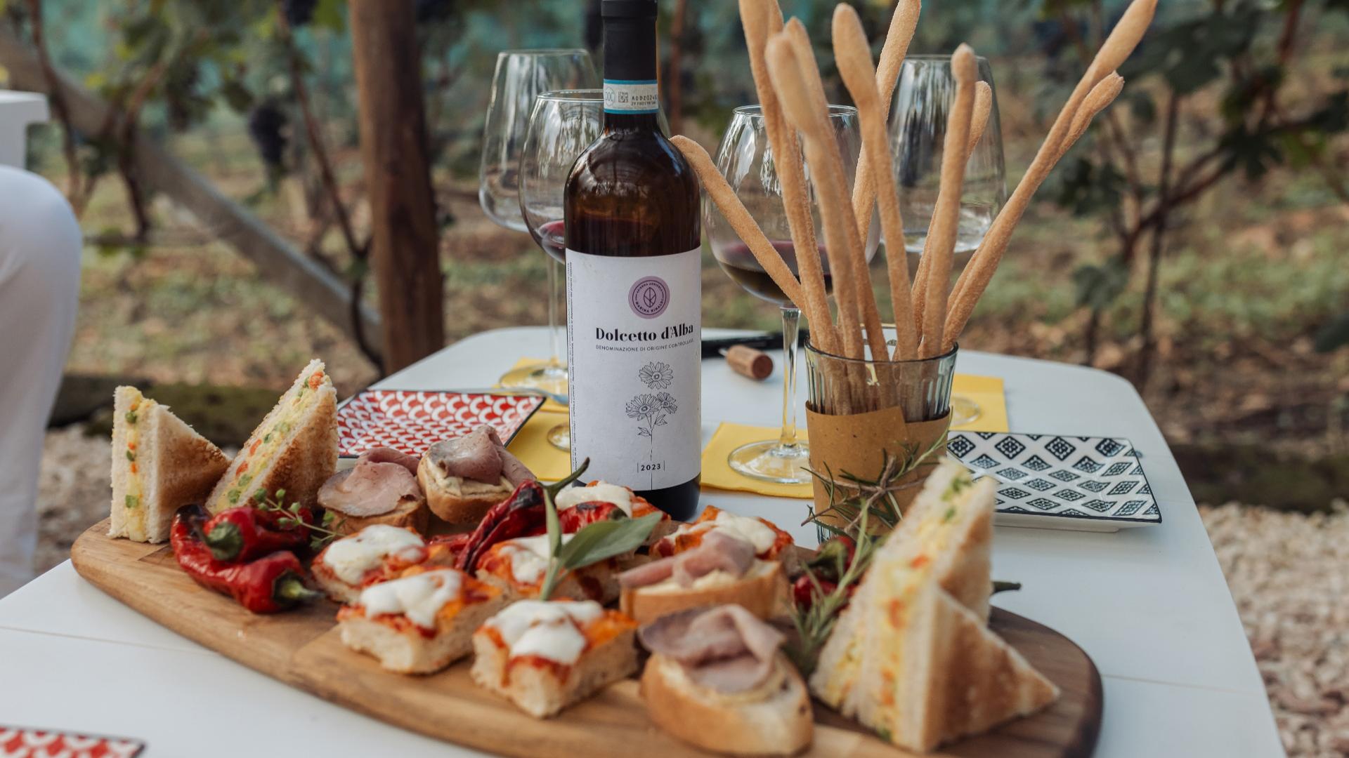 Aperitivo con vino Dolcetto d'Alba, tramezzini e grissini su tavola all'aperto.